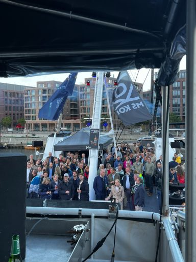 Menschenmenge bei einer Veranstaltung auf dem Wasser mit Flaggen und Gebäuden im Hintergrund.