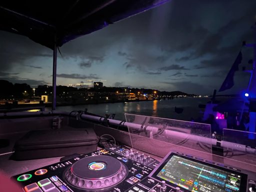 DJ-Setup mit Blick auf eine nächtliche Uferpromenade und beleuchtete Gebäude.